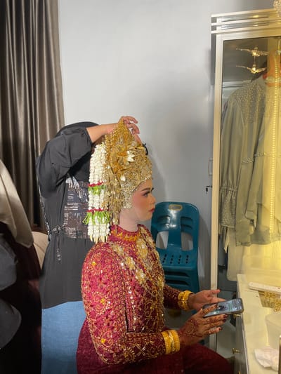 In Tuha Peudaya, Indonesia, a bride adorned in a traditional red and gold wedding gown and elaborate gold headdress is having her hair adjusted by another person. The bride has intricate henna designs on her hands and is holding a mobile phone. The scene takes place indoors, with a closet visible on the right containing clothing, and a teal plastic chair positioned between the bride and the closet. Curtains hang in the background, and the lighting suggests an indoor setting.