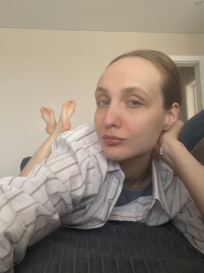 A person lies on a dark gray textured surface, possibly a sofa or bed. They wear a white, vertically striped button-down shirt over a blue undershirt. Their skin appears pale, and they have short, light brown hair. They are wearing a delicate, silver-toned earring with three small, clear stones. Their feet are visible, angled towards the camera. The background consists of a light beige wall and a white door frame. The image is taken at an angle, with the person's head positioned towards the top right corner and their feet toward the bottom left. The location mentioned, Sepych, Russia, does not appear to affect the scene's presentation.