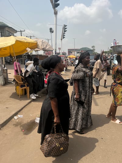 The image presents a busy street scene in Ablekuma, Ghana, under a bright, cloudy sky with distinct shadows cast on the ground, suggesting overhead sunlight. The street surface is a light-colored, dusty pavement, littered with discarded plastic bags and other debris.

In the immediate foreground, a woman wearing a black dress and a dark headwrap is positioned on the left, carrying a brown and tan checkered duffel bag with gold zippers in her right hand. To her right, another woman, also wearing a dark headwrap, stands in a long, leopard-print patterned dress, holding a dark, rectangular object (possibly a phone or wallet) in her right hand. Partially visible on the far right, a third woman in a multi-colored patterned dress walks from right to left, balancing a large, round silver metal basin on her head. The basin contains several clear plastic bottles and white containers. She wears light-colored slip-on sandals.

In the midground, to the left, a yellow open market umbrella and a smaller, closed beige umbrella are visible, sheltering an informal selling area. Beneath the yellow umbrella, a man in a white t-shirt, dark pants, and white sneakers is seated on a beige plastic chair. Another person in a pink top is partially visible behind him. Further into the midground, a group of men stands near parked vehicles; one man in a light-colored t-shirt and shorts gestures upwards with his right hand.

The background reveals a bustling urban environment. A tall pole supporting multiple sets of black traffic lights and loudspeakers dominates the central and right areas. Overhead utility wires are visible. Various vehicles, including a red van and a dark SUV, are either parked or in motion. Buildings with differing architectural styles line the street, one featuring a partially visible red and white "KFC" sign. Market stalls with goods and other individuals populate the far background.