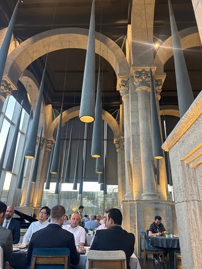 The image captures the interior of a restaurant, likely in Cairo, Egypt, characterized by its grand architecture and unique lighting fixtures. The restaurant features a high ceiling with exposed beams and arched structures reminiscent of Romanesque or Gothic design. Massive stone pillars with ornate capitals support the upper level. Suspended from the ceiling are numerous elongated, cone-shaped pendant lights, casting a warm glow and creating a dramatic visual effect. Large windows line one side of the restaurant, offering a glimpse of the outdoors and letting in natural light.
Several groups of people are seated at tables, engaged in dining and conversation. In the foreground, on the left, a group of men in business attire are seated at a table, with one man facing away from the camera, seemingly in conversation. To the right, another table is occupied by a man in a black patterned shirt, looking down at his phone. Other diners are visible in the background, some appearing to be in formal wear. The tables are set with dark tablecloths and placemats. The overall ambiance suggests a sophisticated dining establishment, possibly a fine dining restaurant or a venue for special occasions. The architectural style and scale indicate a location that aims for a sense of opulence and grandeur.