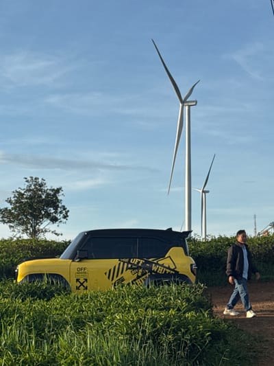 A yellow SUV, adorned with black and white "Off-White" branding, is parked amidst tall green grass. A man wearing a dark jacket, blue shirt, and jeans walks on a dirt path to the right of the vehicle. In the background, two large wind turbines stand against a clear blue sky with a few wispy clouds. A lone tree with dense foliage is visible to the left of the SUV. The scene appears to be in an open, rural area, likely the highlands of Phát Chi, Vietnam, given the presence of wind turbines.