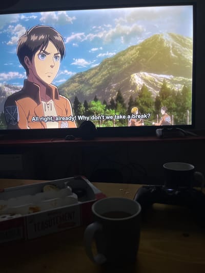 The image depicts an indoor scene in a dimly lit room in Leverkusen, Germany. A large, wall-mounted or stand-supported flat-screen television dominates the upper portion, displaying an anime scene from "Attack on Titan." The central character, Eren Yeager, wearing a brown military-style jacket with the Survey Corps emblem, is visible on the left side of the screen, looking to the right with a determined expression. Subtitles at the bottom of the screen read: "All right, already! Why don't we take a break?" The anime background features a bright blue sky with scattered clouds, verdant mountains, and green trees. Two smaller, similarly uniformed characters are visible in the mid-ground of the anime scene, one appearing to hold a stick or spear.
Beneath the television is a dark wooden shelf or media console, on which a dark, indeterminate object (possibly a remote control) rests. Cables are visible behind this console.
The foreground features a wooden table, partially illuminated, displaying several items. On the left, an open white and red cardboard package labeled "TEARSUTEMENT" contains several individually wrapped items, possibly tea bags or snacks; some appear to have been removed. Two dark grey or black ceramic mugs are present on the table. The mug closer to the viewer contains a dark liquid. A black PlayStation game controller is positioned to the right of the package, behind the closer mug, with a second dark mug behind it. Small, metallic, circular objects are scattered on the table between the package and the controller. The overall impression is of a casual, relaxed setting, likely during evening hours, with an individual engaging in media consumption and refreshments.