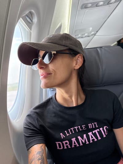A woman is sitting in an airplane seat, looking out the window. She is wearing a baseball cap, sunglasses, and a black t-shirt with the words "A LITTLE BIT DRAMATIC" printed on it in pink. She has tattoos on her arm. The interior of the airplane is visible, including the overhead compartments and seats. The woman's face is turned to the left, and she is smiling slightly.