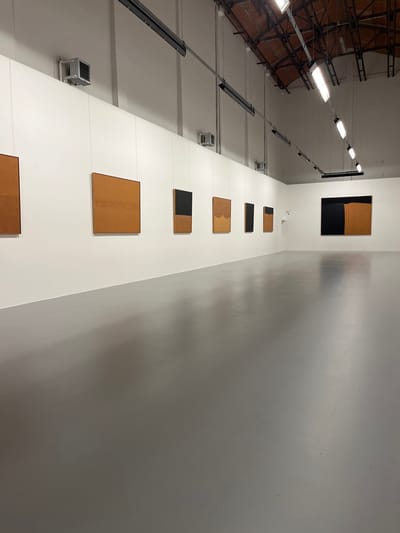 The image captures an interior view of a large, high-ceilinged space, likely an art gallery or exhibition hall. The perspective is looking upwards along a tall, white wall that serves as a display surface for a series of artworks.
On the left side of the frame, several prominent overhead light fixtures are visible, extending in a linear fashion across the high ceiling. These fixtures appear to be long, rectangular units with multiple bright bulbs providing ample illumination. Below these lights, a darker, exposed structural ceiling or series of beams can be seen, hinting at an industrial or minimalist architectural style. Further down the white wall, several square-shaped vents or smaller lighting units are mounted.
The main focal point is the vertical arrangement of seven rectangular artworks displayed along the pristine white wall. These pieces primarily feature an earthy, light brown or tan color, possibly resembling wood, cork, or a natural fabric, often contrasted with sections of solid black.
Starting from the top and moving downwards:
1. The topmost visible artwork is a large, tall rectangle, predominantly tan with a significant black section at the bottom.
2. Below it is a smaller, horizontal rectangular piece, also tan with a black bottom section.
3. The third piece is a small, horizontal rectangle, entirely black.
4. The fourth artwork is a medium-sized, horizontal tan rectangle with a subtly curved bottom edge, giving it a slightly organic shape.
5. Following that is another medium, horizontal tan rectangle with a black section covering its right side.
6. The sixth piece is a larger, squarish tan rectangle, featuring subtle vertical textural lines or markings.
7. The seventh and lowest visible artwork is a horizontal tan rectangle, partially cropped at the bottom of the frame.
All artworks are hung with thin, almost invisible wires, maintaining a clean and minimalist presentation against the white background. To the right of the wall displaying the art, another wall appears in a lighter gray tone, reflecting some ambient light and exhibiting subtle shadows and texture, suggesting a wider space. The overall impression is one of modern aesthetics, focusing on form, color, and texture within a well-lit, expansive environment. No people or visible text are present in the image.