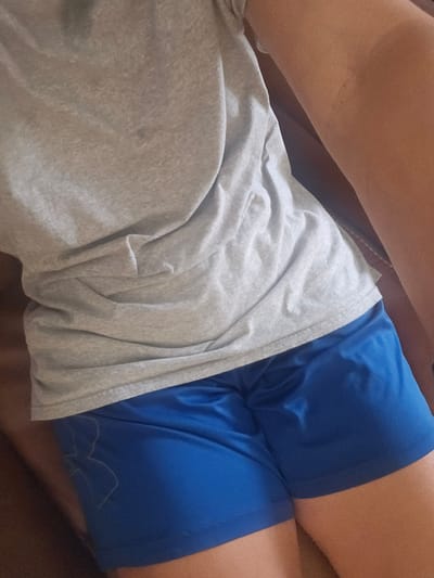 This is a close-up, medium-shot, eye-level photograph of a person from the waist down, likely an adult, wearing a light gray t-shirt and bright blue athletic shorts. The t-shirt is creased and somewhat rumpled, with a dark speck visible on the fabric. The blue shorts appear to be made of a synthetic material and have a subtle sheen. An outline of a logo, possibly an athletic brand, is visible in white on the left thigh of the shorts. The person is positioned on a brown surface, possibly furniture. The lighting suggests an indoor environment. There is no indication of the city of Islamorada, United States of America, or any other specific location.