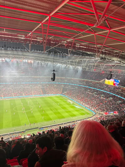 The image is taken from a high vantage point inside Wembley Stadium, located in Wembley, United Kingdom, at coordinates 51.558 latitude and -0.28 longitude. The image captures a football match in progress on the pitch below. The stadium is packed with spectators, many wearing red and white. The pitch is green, and a light blue scoreboard is visible in the background. The image shows a group of people in the stands, including a woman with blonde hair, a man with dark hair, and a group of people in the background. The image shows the stadium's roof, which is a complex metal structure with red beams. The stadium lights are on, illuminating the pitch. The image captures the atmosphere of a football match.