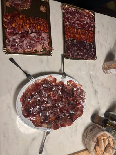 The image shows a table laden with Spanish tapas. There are two rectangular trays, one with three sections of cured meats (likely chorizo and jamón), and another with two. A large plate is piled high with glistening slices of jamón, accompanied by three forks. A bottle of rosé wine, small beer bottles, and a basket of bread are also present. The marble tabletop adds a refined touch to the presentation.