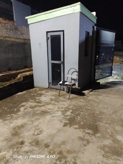 The image depicts a small, grey, rectangular structure, possibly a guardhouse or small office, situated outdoors. The building has a light green trim along its roofline. A glass-paned door with a dark frame is visible on the front of the structure. To the right of the building, a black metal gate with a decorative pattern is partially open. In front of the door, a metal chair with a light-colored cushion and what appears to be a book or papers on it is placed on a concrete surface. The ground is a weathered concrete pavement with some discolored patches, suggesting it might be wet or dirty. Behind the structure, there is a low, rough-hewn wall or embankment made of rocks and soil. The background is dark, indicating the image was taken at night or in low light conditions. There is a visible watermark in the bottom left corner that reads "Shot on AWESOME A90."
The image was taken in Ibadan, Nigeria, based on the watermark, which is a common feature on photos taken with certain mobile devices. The scene appears to be an outdoor area adjacent to a property, likely a residential or commercial compound, given the presence of the gate and the structure that could serve as a security post. The lighting suggests artificial light sources are present, possibly from the building itself or nearby streetlights, casting shadows on the ground. There are no people visible in the frame. The overall impression is of a functional, utilitarian space, captured in a somewhat stark and unadorned manner.