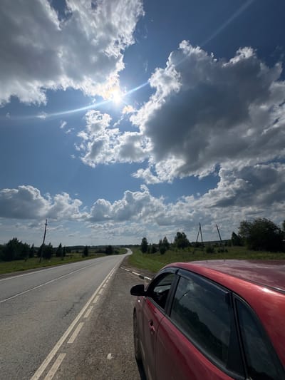 This is a vertical, eye-level shot of a red car parked on the shoulder of a paved road. The road curves to the left and disappears into the distance. The sky is a mix of bright blue and dark, ominous clouds, with the sun peeking through the clouds and creating a lens flare effect. The landscape on either side of the road is a flat, grassy field dotted with trees. There are utility poles visible in the distance. The image appears to have been taken during the day in a rural area. The location is identified as Rozhdestvensk, Russia.