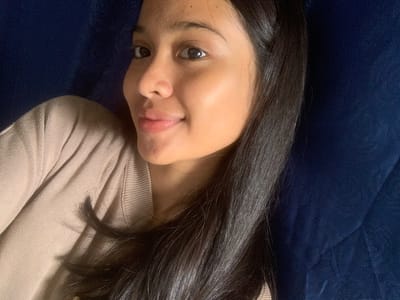 A close-up, overhead shot shows a young woman with dark hair and brown eyes lying on a dark blue surface, possibly a couch or bed. She is wearing a tan, ribbed sweater. Her head is turned to the right, and she is looking towards the camera with a slight smile. Her skin is clear, and her lips are tinted pink. The lighting suggests an indoor environment, with soft illumination highlighting her face and the texture of her clothing. There are no visible contextual clues in the image that would indicate the specific location within Sungai Meriam, Indonesia.