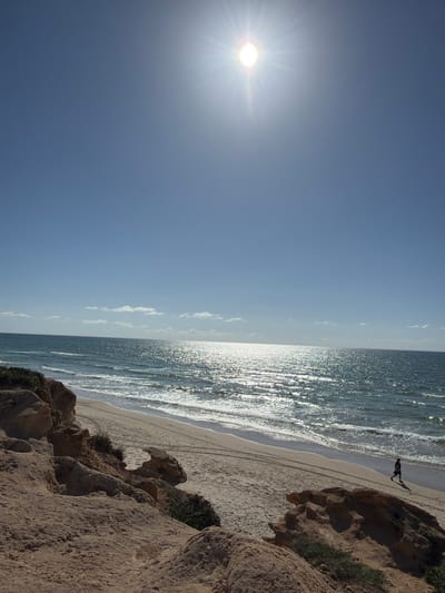 The image displays a sunny coastal landscape at Beit Yanai, Israel. The upper portion of the frame features a clear blue sky with the sun, appearing as a bright, starburst-like disc, positioned in the upper right quadrant. The Mediterranean Sea spans the mid-ground, its surface highly reflective from the sun, creating a shimmering path of light towards the viewer. Gentle waves break onto a light-colored sandy beach. A solitary figure, dressed in dark clothing, is observed walking from right to left along the water's edge on the sand. Parallel tire tracks are visible on the beach sand. The foreground and left side of the image consist of eroded, reddish-brown sandstone cliffs with sparse green vegetation growing on and between the rock formations.