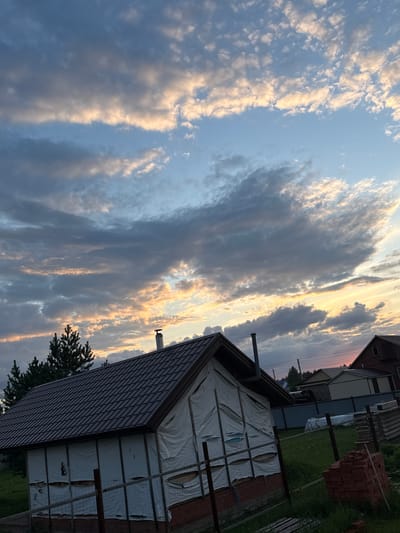 The image depicts a rural or semi-rural scene under a dramatic sky, likely at dusk, in Nizhnyarkeyevo, Russia. In the foreground, a building under construction is visible, with its walls covered in white protective sheeting that is partially torn and revealing wooden framing and insulation. The building has a dark brown metal roof with a chimney. To the left of this structure, a dark green pine tree is prominent. In the background, several other buildings and structures can be seen, including what appears to be a house with a red roof. The sky is filled with a mix of dark and light clouds, with streaks of orange and yellow light indicating the setting sun. The ground is covered in green grass and appears somewhat overgrown in places. A pile of red bricks is stacked to the right of the frame, suggesting ongoing construction or renovation work. A wire fence runs across the midground. The overall impression is one of a quiet, developing settlement under a visually striking sky.