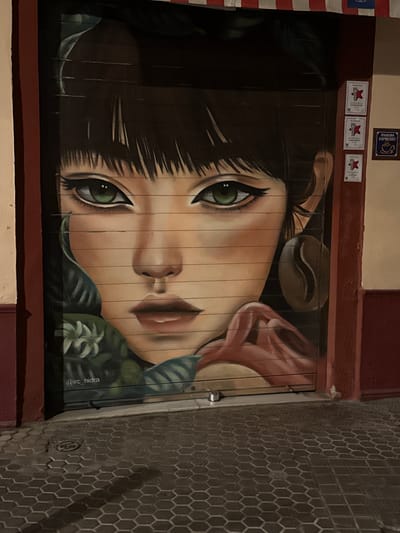 The image is a photograph of a mural painted on a wooden surface, likely a door or shutter, in Seville, Spain. The mural features a close-up, stylized portrait of a young woman's face, depicted with large, striking green eyes and dark hair. Her head is tilted, giving her a contemplative or perhaps wistful expression. A single coffee bean is artfully placed above her head, and elements of lush green foliage frame her face, suggesting a connection to coffee cultivation or a natural, organic theme. The mural is rendered with smooth, blended colors and bold lines, giving it a graphic and illustrative quality.
Above the mural, on the wall, are several framed certificates and a small sign. One sign is partially visible and appears to say "IPANEMA ESPRESSO" with a coffee cup logo. The certificates have a red star symbol, indicating some form of recognition or award.
To the right of the mural is a textured wall covered in hexagonal tiles, common in Spanish architecture. The lighting suggests it is nighttime or dusk, as the scene is illuminated by artificial light, casting shadows on the tiled wall. The overall impression is of a charming, artistic facade of a cafe or business, integrated into the urban landscape of Seville. The visible text on the mural itself, "@rc_hidra", is likely the artist's signature or social media handle.