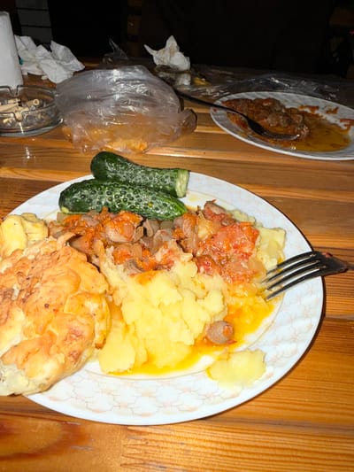 The image shows a plate of food on a wooden table. The plate contains mashed potatoes, a fried cutlet, stewed meat with tomatoes and onions, and two cucumbers. There is also a fork on the plate. In the background, there is an ashtray with cigarette butts and some plastic bags. The lighting suggests the photo was taken at night or in a dimly lit indoor setting. The overall scene appears to be a casual meal, likely at a home or outdoor seating area.