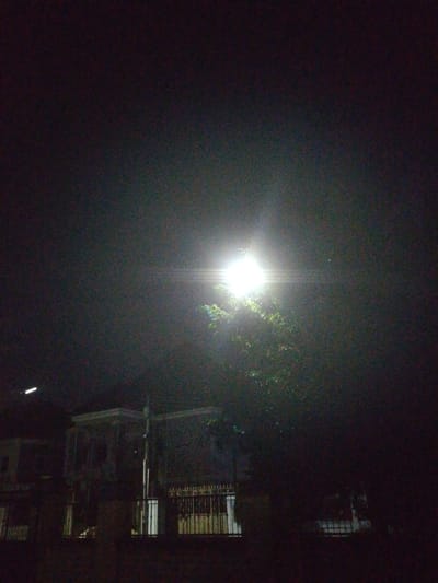 An outdoor, nighttime, low-light, wide-angle shot captures a streetlamp illuminating a tree and the facade of a two-story building. The streetlamp is positioned to the right of the image's center, casting a bright, diffused light that creates a lens flare effect. The tree, partially lit by the lamp, is dense with foliage and appears to be in front of the building. The building, located in Rumuomasi, Nigeria, exhibits an architectural style with visible windows and a veranda-like structure on the upper level. In the foreground, a metal fence runs across the lower portion of the image. The overall environment is dark, with the illumination from the streetlamp being the primary light source. There are no people visible in the scene.