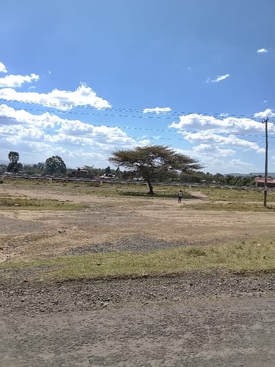 The image depicts an expansive, open landscape in the Kiambu Township ward, Kenya, under a bright blue sky with scattered white cumulus clouds. The foreground consists of a gravel and dirt surface with interspersed dry grass. The midground features a vast, sparsely vegetated field of light brown soil and patches of green grass. A single, mature tree with a wide, flat, green-brown canopy stands prominently to the left of the center. One individual, wearing a light-colored top and dark bottoms, walks away from the viewer on a dirt path in the midground, positioned slightly to the right of the large tree. In the background, several distant buildings, some with discernible red roofs, are visible amidst additional trees and a horizontal white pipeline or barrier structure. A dark utility pole supporting multiple overhead power lines is present on the far right side of the frame, with more power lines stretching horizontally across the upper portion of the sky.