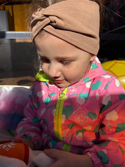 A close-up shot captures a young child with fair skin, appearing to be asleep. The child is wearing a light brown head covering and a pink and green patterned jacket. The jacket features images of white clouds and green hearts, with the word "Outdoor" visible. Part of a yellow balloon is visible in the background. The image seems to have been taken in Кипчак, Russia.