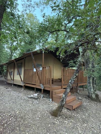 A canvas glamping tent with wooden decking and steps is situated in a wooded area. The tent is beige with a dark brown roof and is supported by visible poles. A wooden railing encloses the deck, and a sign with the number "9" is visible on the railing. The ground around the tent is covered with gravel and fallen leaves. The scene is surrounded by lush green trees, with some branches extending over the tent and deck. A light blue sky is visible through the leaves at the top of the image. The glamping tent is located in Cavriglia, Italy.