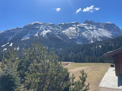 The image depicts a mountainous landscape, likely in the vicinity of Sappada, Italy, considering the location information provided. A prominent mountain range with snow-capped peaks dominates the background. A dense forest covers the lower slopes of the mountains, transitioning to a grassy field in the foreground. A structure, possibly a chalet or cabin, is partially visible on the right side of the frame, with a stone-paved terrace or walkway. In the foreground, pine trees are visible. The sky is blue with a few scattered clouds and a visible sun glare. The overall scene suggests a sunny day in a mountain resort or rural setting. There are no people visible in the image.