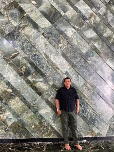 In Aluekambuk, Indonesia, a man stands barefoot in front of a wall adorned with diagonal green and gray marble tiles. He is wearing a black short-sleeved collared shirt and dark green pants, with his hands in his pockets. A watch is visible on his left wrist. The floor is also tiled with dark marble.