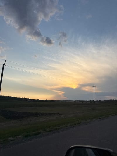 The image captures a rural landscape in Sadaclia, Moldova, during what appears to be sunset. The sky is a prominent feature, displaying a gradient of colors from a pale blue at the top, transitioning through soft yellows and oranges near the horizon, with scattered clouds of varying shades of gray and white. Wispy, elongated clouds are visible, suggesting wind movement.

In the foreground, a paved road runs diagonally from the bottom right corner, leading into the distance. Along the left side of the road, there is a dark, uneven terrain with patches of green grass. Further into the midground, a dark, cultivated field is visible, with a distinct darker patch that could indicate tilled soil or a different crop. Beyond this field, a gently rolling landscape with distant trees and possibly some structures can be seen against the horizon.

Two utility poles with wires are present in the image. One is positioned on the far left, a tall, dark pole with wires extending horizontally across the frame. The second pole is on the right side, also dark and appearing to be of similar construction, with wires also running horizontally. The overall lighting suggests it is either early morning or late evening, with the sun partially obscured by clouds near the horizon. There are no people or animals visible in the scene. The atmosphere is serene and natural, indicative of a rural setting.