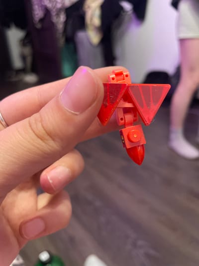 A close-up, eye-level shot shows a person's hand holding a small, red, multi-jointed toy. The toy has triangular red wing-like attachments and a pointed red tip. The hand has pale skin, short fingernails, and a silver ring on the index finger. In the blurred background, a person's legs are visible, wearing white socks and a light-colored skirt or shorts. The setting appears to be indoors, with a wooden floor and a dark, cluttered background possibly indicating a closet or room with clothes. The image does not provide any information about the city of Borisova Griva, Russia.
