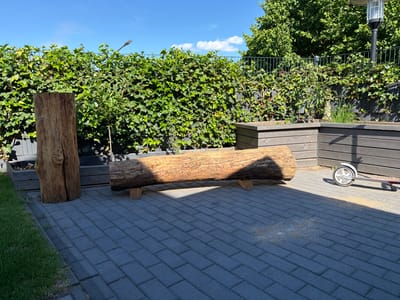 This is a photograph taken on a sunny day, likely in Bissendorf, Germany, depicting an outdoor paved area adjacent to a lush green hedge. The paving stones are a dark grey rectangular brick pattern. To the left, a tall, rough-hewn wooden log stands upright. In the center of the paved area, a long, thick log is placed horizontally on two smaller wooden supports, forming a natural bench. Behind the logs, a dense green hedge grows along a dark fence. In the background, to the right, a dark wooden planter box is visible, and next to it, a child's scooter with two wheels. The sky above is a clear blue with a few scattered white clouds. The lighting suggests it is mid-day, with strong shadows cast by the objects on the paving stones.
