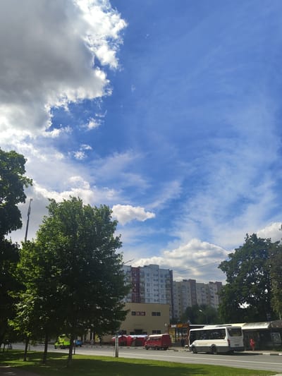 This media file depicts a street scene in Ramenskoye, Russia. The image is oriented vertically, showing a wide expanse of blue sky with scattered white clouds. Tall apartment buildings, typical of Soviet-era architecture, line the street on the left. A white bus with yellow trim and a red truck are visible on the road, suggesting public transportation and commercial activity. The presence of green trees and a grassy area indicates an urban park or residential area with landscaping. The lighting suggests it is daytime, with the sun casting shadows and illuminating the clouds. No specific activity or event is clearly depicted, but the scene appears to be a typical day in a Russian city. The weather seems pleasant, with a mix of sun and clouds. Some text is visible on the side of the bus and on signs in front of the buildings, but it is not legible in detail.