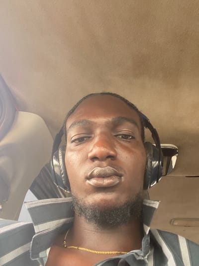 A man is indoors, wearing headphones and a striped shirt with a gold chain. The location is presumed to be inside a car, looking up at the ceiling. The specific location in the city of Odo Ona, Nigeria is not determinable from this image.