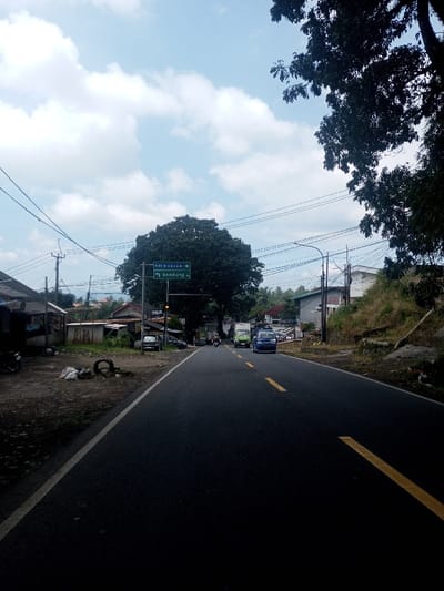 The scene depicts a daytime street view in Niz Lakuk, India, focusing on a two-lane asphalt road. The road features a solid yellow median line transitioning into a dashed yellow line in the distance, indicating two-way traffic. On the immediate left, a dirt shoulder is present, littered with white sacks and two dark tires. A dark motorcycle is parked on this shoulder. Beyond, a series of modest, single-story buildings with various roof types, including corrugated metal, line the roadside. Overhead, a network of power lines spans across utility poles. A green road sign, affixed to a pole, indicates directions for "PIM. R. Gumuruh" to the left and "Bandung" with a right-turn arrow. Further down the left side, dark-colored cars are parked.

On the right side of the road, an unkempt grassy embankment rises. A white building with a dark roof is situated on this higher ground. A street light pole with a yellow fixture is visible near the embankment. Dark green tree foliage partially obscures the upper right sky. Overhead power lines are also visible on this side.

Vehicular traffic is present on the road, moving away from the viewer. A light blue microvan or minibus occupies the right lane, followed by a green microvan or minibus in the same lane. Further ahead, a white truck and other indistinct vehicles, including at least two motorcycles, are visible. The sky is partially cloudy with white cumulus clouds against a light blue background. The overall lighting suggests bright daylight conditions. No distinct individuals are clearly visible.