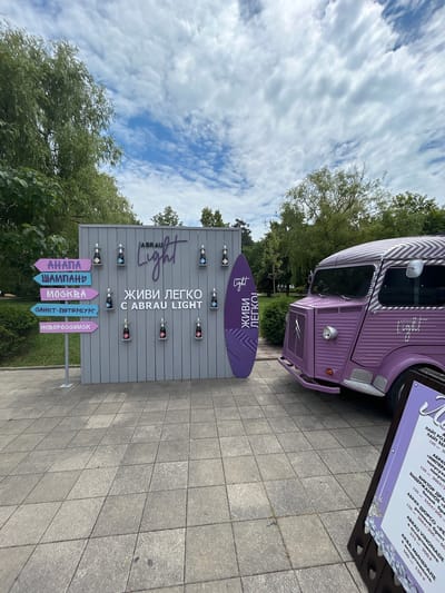 This media file captures an outdoor scene featuring a purple vintage Citroën van with a striped roof and a large, grey sign shaped like a surfboard. The sign is adorned with decorative bottle shelves, some of which hold small bottles. The text on the surfboard-shaped sign reads "ABRAU Light" and "ЖИВИ ЛЕГКО C ABRAU LIGHT" (Live Lightly with Abrau Light). Below this, a series of directional arrows point to different Russian cities: "АНАПА" (Anapa), "ШАМПАНЬ" (Champagne), "МОСКВА" (Moscow), "САНКТ-ПЕТЕРБУРГ" (Saint Petersburg), and "НОВОРОССИИСК" (Novorossiysk).
Another sign, partially visible to the right of the van, displays a menu with text in Russian, including wine names like "ЛАЙТ РОЗ" (Light Rose), "АБРАУ «ИМПЕРАТОР»" (Abrau "Emperor"), and "АБРАУ-ДЮРСО «РЕЗЕРВ»" (Abrau-Durso "Reserve"), along with prices. The setting appears to be a paved area, possibly a promenade or an event space, with greenery and trees in the background. The sky is cloudy, suggesting it might be daytime, but the lighting is somewhat diffused. The overall impression is of a promotional event or a branded installation, likely associated with the Abrau-Durso winery. The scene is static, with no people visible.