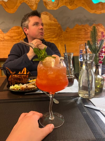 A restaurant table features a large cocktail in a stemmed glass, garnished with mint and fruit, accompanied by a small basket of fries and spring rolls. Nearby, there are decorative glass bottles and foliage. A man is seated at the table, looking towards the cocktail with his hands clasped. A person's hand touches the base of the glass on the right. The table is covered with a textured black mat. The background includes a patterned wall.