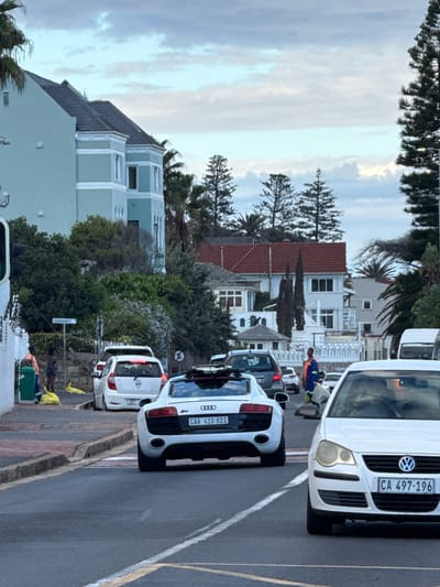 The image shows a street scene in Muizenberg, South Africa. Several cars are lined up on the street, including a white Volkswagen Polo with the license plate "CA 497-196" and a white Audi with the license plate "CAA 410-823". A person wearing a blue and orange work outfit is standing near the cars. Houses and trees are visible in the background. A street sign reading "SAMPBURST" is also visible. The sky is cloudy.