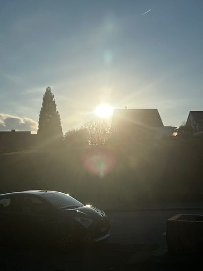 The image depicts a static outdoor scene in a residential area of Rafz, Switzerland, under strong backlighting conditions, suggesting either early morning or late afternoon. The sky is predominantly clear blue with faint wisps of clouds visible on the left side and a distinct white contrail stretching diagonally across the upper-right quadrant. The sun is a prominent, bright orb positioned centrally in the upper-midground, casting intense glare and multiple lens flares, including circular and polygonal artifacts, across the frame.

In the foreground, a dark-colored compact hatchback car, possibly parked, is visible on the left, angled slightly towards the right. Its front section and windshield display reflections from the sun, while the remainder of the vehicle appears in silhouette. To the right of the car, a dark, rectangular concrete planter or basin is situated on what appears to be a paved surface.

The background features a gently rising grassy slope behind the paved area. On this elevated ground, several residential buildings with dark, sloped roofs are partially obscured by the intense sunlight and the silhouetted terrain. A tall, conical evergreen tree stands prominently on the left side of the background, contrasting with several bare deciduous trees to its right. No individuals or specific actions are discernible within the scene.