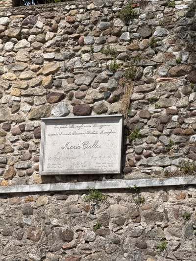 The image shows a weathered stone wall in Sirmione, Italy, with a plaque commemorating Maria Callas. The plaque, set into the wall, is made of a lighter stone and features an inscription in Italian. The text appears to be in a script font, with the name "Maria Callas" prominently displayed in larger lettering. Below this, there are dates and possibly other biographical details. The wall itself is constructed from irregular, variously colored stones, typical of historical architecture. Some small plants and dry vegetation are growing in the crevices of the wall, suggesting age and exposure to the elements. The scene is illuminated by natural light, casting shadows that highlight the textures of the stones and the relief of the inscription.