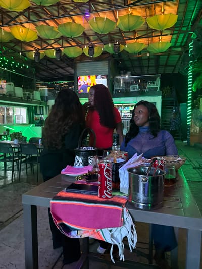 The image captures an evening scene at a leisure establishment in Falomo, Nigeria. Three women are present around a gray rectangular table. The woman on the left has her back to the camera, revealing long, dark, curly hair and a dark top. The central woman, also with long, dark, curly hair, wears a red top and has her mouth open, indicative of speaking or singing. The woman on the right, with long, dark, wavy hair, wears a sheer blue-purple long-sleeved top over a dark bra and is looking to her left.

The table holds a colorful, striped fabric runner, a black smartphone, a red Coca-Cola can, a bottle of Jack Daniel's whiskey, a white napkin, a metal ice bucket with tongs, a glass containing amber liquid and ice, a woven basket, a black and white patterned handbag, a pink fabric, and a string of beads.

The establishment's ceiling features exposed metal beams from which numerous inverted, yellow-green umbrellas hang as decorative elements, along with spotlights. Green LED strip lights illuminate architectural features and foliage throughout the space. In the background, a large television screen displays colorful content. A bar area with bottles and glassware is visible, alongside additional tables and chairs. A staircase with white railings is present in the far right background, with a person ascending or descending. The overall lighting scheme is predominantly green, creating a distinct ambiance.