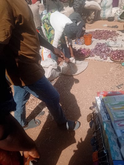 The scene depicts an outdoor market in Gurku Gwari, Nigeria, under bright sunlight, casting prominent shadows. The ground is a light-brown dirt or sandy surface. In the immediate foreground, the lower torso and legs of an individual wearing a brown shirt, blue jeans, and grey sandals are visible, with their right hand extended towards a small, light-colored fabric bag. Centrally, a person wearing a white long-sleeved top and a multi-patterned skirt (green, yellow, black design) is bent at the waist, reaching into or handling this same fabric bag. This individual has dark hair. Surrounding this interaction are multiple displays of red onions, arranged in piles directly on the ground, on a mat, and within woven baskets. An orange plastic basket is also present among the onions. In the background, several large, light-colored woven sacks, some marked "KANG," are stacked. Further to the right, a vertical display rack holds numerous packaged items, appearing as consumer goods. Additional, partially obscured individuals are visible in the background, engaged in various market activities.
