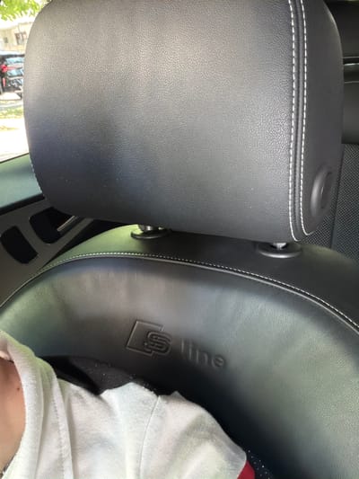 The image shows a close-up view of the interior of a car, specifically focusing on black leather car seats. The seat on the left appears to be a headrest, with white contrast stitching along its edge. It is positioned next to a car seat on the right, which has a subtle embossed logo that appears to be "S line". A white fabric, possibly a shirt or towel, is draped over the right seat, and a person's shoulder and arm are visible beneath it. The overall impression is that of a parked car on a bright day, as indicated by the natural light illuminating the scene. There are no overt signs of activity, suggesting the car is stationary. The focus is on the material and branding of the car's interior. The location context provided is Orhei, Moldova.