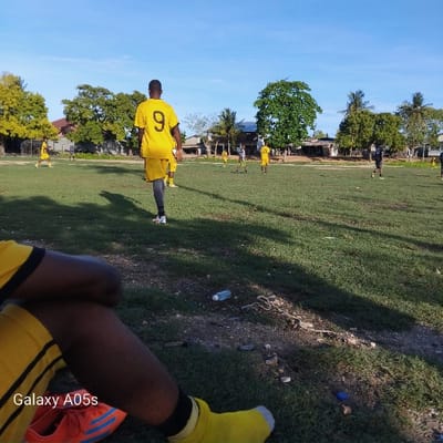 A grassy soccer field in Nungwi, Tanzania, is the scene for a game of soccer between two teams of young men. The player wearing the number 9 on the yellow jersey is looking away from the camera and towards his team. A player in a yellow jersey is walking in the distance. The soccer field is bordered by trees on either side. In the foreground, a person wearing yellow socks and a black and yellow jersey is sitting with their legs crossed and facing the camera. The person has a black Galaxy A05s phone in their left hand. The phone is resting on the grass with a soccer shoe visible in the foreground. The surface of the field is green grass with patches of dirt and small rocks.
