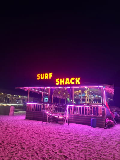 At night in Arambol, India, a multi-tiered wooden structure stands next to a beach. The building is illuminated with purple neon lights that also cast a purple hue on the sand. The words "SURF SHACK" are vertically displayed in bright yellow neon. The building has multiple levels with wooden railings and staircases. People can be seen on one of the levels. The beach extends to the right of the building.