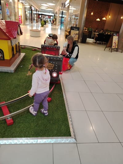 A young girl with pigtails, wearing a pink hoodie and purple pants, stands on a grassy area in front of a toy train. The train, which appears to be part of a play area, has a sign that reads "WESTERN TRAIN". Another child, a boy, sits in the train, facing away from the camera. A woman in a black vest and jeans is crouched nearby, looking at her purse. In the background, the scene is set in a shopping mall. Shops with displays are visible, along with mall seating areas and a cafe with patrons. The floor is tiled, and lighting fixtures are visible on the ceiling. A cafe with a rustic design featuring wooden elements and pendant lights is also present. Signs in Polish, such as "Wołomin Smaki" (Wołomin Tastes), are visible, indicating the location as Wołomin, Poland. The overall atmosphere suggests a daytime scene in a public indoor space.