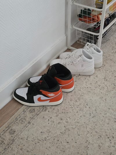 In Montrouge, France, inside a dwelling, a low-angle shot captures a pair of black, white, and orange high-top sneakers resting against a white baseboard on a patterned rug. To the right of these sneakers, a white shoe rack with two wire baskets is visible. The top basket contains folded clothing in orange, green, and off-white hues, alongside yellow and white packaging. The bottom basket holds a grey cylindrical object, possibly a vacuum cleaner attachment, and other dark items. Behind these sneakers and to the left, another pair of white low-top sneakers is positioned. The overall scene depicts an organized storage area for footwear and other household items.