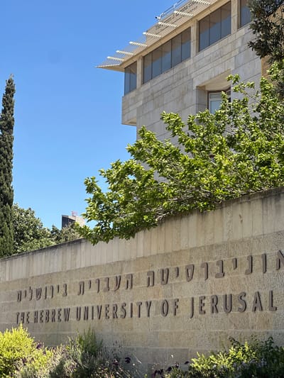 The image shows a building with the text "THE HEBREW UNIVERSITY OF JERUSALEM" carved into its stone facade. The text is in both English and Hebrew. The building appears to be made of light-colored stone blocks. Green trees and foliage are growing around the building, some branches hanging over the text. The sky is clear and blue, suggesting a sunny day. The perspective is from below, looking up at the building and the sky. A portion of another building with windows and a metal railing is visible on the left. The overall impression is of a university campus in a warm climate.