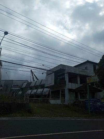 A low-angle shot captures a weathered, two-story building under a cloudy sky, intertwined with numerous overhead power lines. Adjacent to the building is a sloping roof structure with repeating angular patterns. A small gazebo-like structure is attached to the building's right side. A road stretches across the bottom of the image, suggesting an urban or suburban setting. The overall tone is muted due to the overcast conditions.