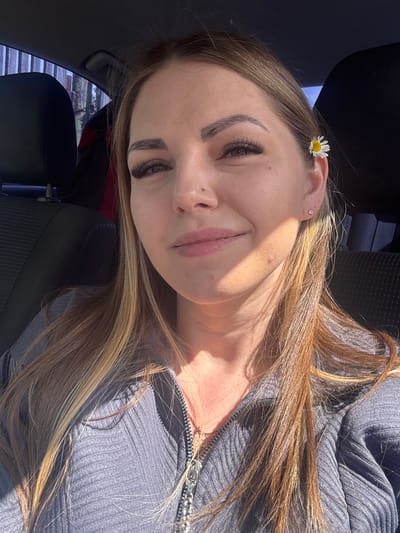 A young woman is sitting in what appears to be a car, based on the seat and interior details visible. She is angled to the camera, displaying a close-up view of her face and upper body. She is wearing a grey ribbed top, partially unzipped, revealing a hint of a necklace. Her light brown hair, with blonde highlights, falls across her shoulder and around her face. A single daisy is tucked behind her ear. The lighting is bright, casting shadows across her face. The location cannot be determined from the given image; there is no indication of Slobodka, Russia.