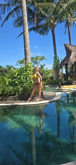 In Tibubiyu, Indonesia, a light-skinned adult female with light-colored hair, wearing a yellow bikini top and black bikini bottoms, stands barefoot on the edge of a swimming pool. She faces the viewer, smiling, with her hands on her hips, her right leg slightly bent. Her reflection, along with that of two prominent palm trees, is visible in the turquoise water of the pool. Behind her is a dense, lush green hedge or bush, bordered by a dark, possibly pebbled, retaining wall. Several tall palm trees with green fronds rise into a clear blue sky with minimal cloud cover. To the right, a traditional thatched-roof structure and a dark, possibly stone, statue are visible near the far end of the pool. The scene is illuminated by bright daylight, indicating an outdoor tropical environment.