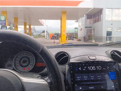 From the driver's seat, a clear view of a Shell gas station is visible. The gas station has multiple yellow support columns and an orange-white canopy. Adjacent to the gas pumps is a modern, two-story building. The car's dashboard features a steering wheel with red stitching, an analog speedometer, and a modern infotainment system displaying a radio frequency of 106.10 MHz.