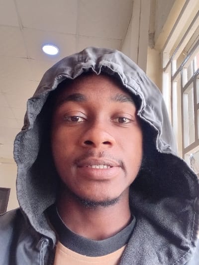 A close-up, frontal portrait depicts a male subject of African descent, centered within the frame. He is wearing a dark grey or black hooded sweatshirt, with the hood pulled up around his head. A light brown shirt with a dark collar is visible beneath the hooded garment. The subject has dark eyes, short dark hair, and a thin goatee extending along his jawline and a mustache. His expression is neutral to slightly smiling, with his mouth slightly open, revealing some upper teeth. The background indicates an indoor environment. To the subject's left, a light-colored wall is visible, with a dark, vertical element partially present in the lower left corner. To the subject's right, a window with horizontal and vertical metal bars (grille) is visible. Above, the ceiling is composed of light-colored grid panels, and a single, round, illuminated white LED light fixture is embedded in the ceiling. The visual information within the image does not provide specific details to confirm the location as Kwazailewa, Nigeria.