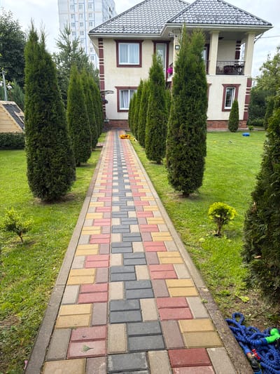 The image depicts a residential property with a paved walkway leading to a two-story house. The walkway is made of multi-colored interlocking pavers in shades of pink, yellow, and gray, laid in a running bond pattern. Flanking the walkway are rows of tall, conical evergreen trees, likely thuja, creating a formal garden effect. The lawn is neatly mowed and appears well-maintained.
In the background, a modern, multi-story building with a light-colored facade and dark-framed windows is visible. The house itself has a beige stucco exterior with dark red trim around the windows and decorative elements. It features a covered balcony with a wrought-iron railing on the second floor.
On the lawn near the house, a blue plastic object, possibly a children's toy, is visible. To the right of the walkway, a coiled blue garden hose with a green nozzle and red valve is present on the grass.
The scene suggests a suburban or semi-rural environment, potentially on the outskirts of a city. The presence of a modern high-rise building in the background could indicate proximity to an urban center. While no people are visible in the image, the presence of toys and gardening equipment suggests a lived-in residential setting. The overall impression is one of a well-kept property. The scene is consistent with a residential area in Solnechnogorsk, Russia, given the architectural style and landscape common in such locations.
