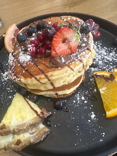 A close-up, high-angle view captures a stack of three golden-brown pancakes presented on a matte black circular plate. The pancakes are topped with a layer of brown chocolate spread, a halved red strawberry, several dark blue blueberries, and clusters of red currants with small green leaves. White powdered sugar is liberally dusted over the pancakes and plate, and dark chocolate syrup is drizzled across the dish. Surrounding the pancake stack on the plate are additional fruit garnishes: a wedge of pineapple with a dark syrup drizzle in the lower left, a slice of orange in the middle right, and a slice of pink grapefruit partially visible in the upper left. The background is blurred, revealing a light brown wooden table surface and a partial clear glass in the upper left corner. The scene depicts a prepared meal, likely breakfast or dessert, situated in the city of Weißenburg in Bayern, Germany.